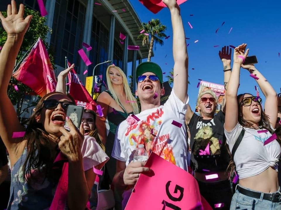 Gruppe fröhlicher Menschen feiert mit Hochhalten von Plakaten und rosa Konfetti.