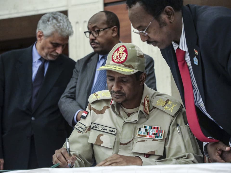 A man signing a document surrounded by three men in military uniform.