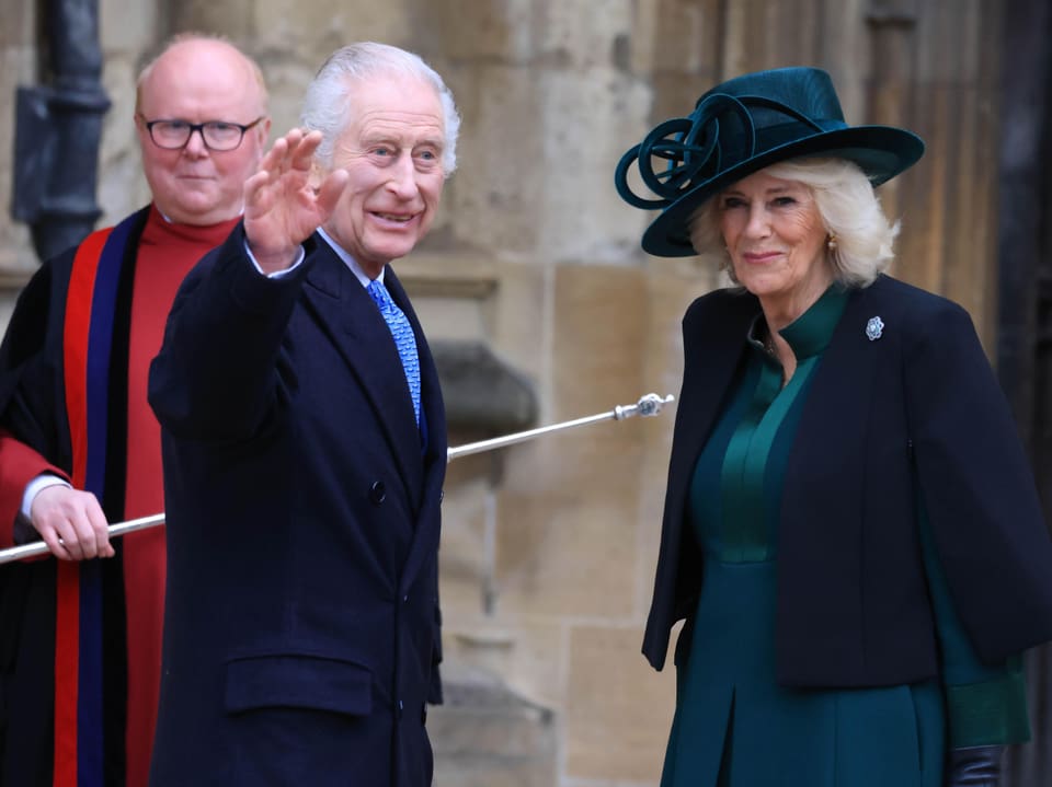 König Charles und seine Frau Königin Camilla stehen vor einer Kirche und winken Menschen zu.