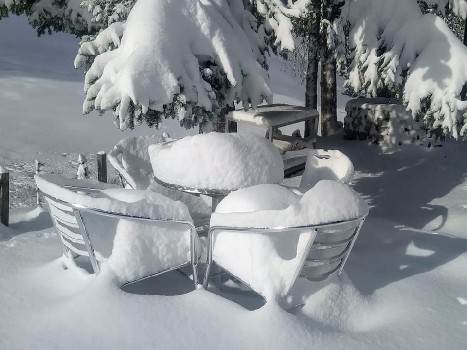 Tisch und Stühle im Freien, bedeckt mit dicker Schneeschicht, umgeben von verschneiten Bäumen.