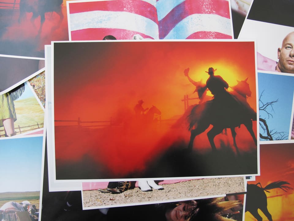 Auf vielen kleinen Abzügen liegt ein Foto von einem Cowboy bei der Arbeit vor feuerrotem Abendhimmel. 