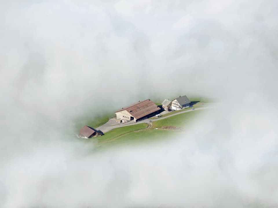Weisses Nebelmeer von oben, in der Mitte ein Loch, durch welches Mann einen Bauernhof und etwas Wiese erkennt.