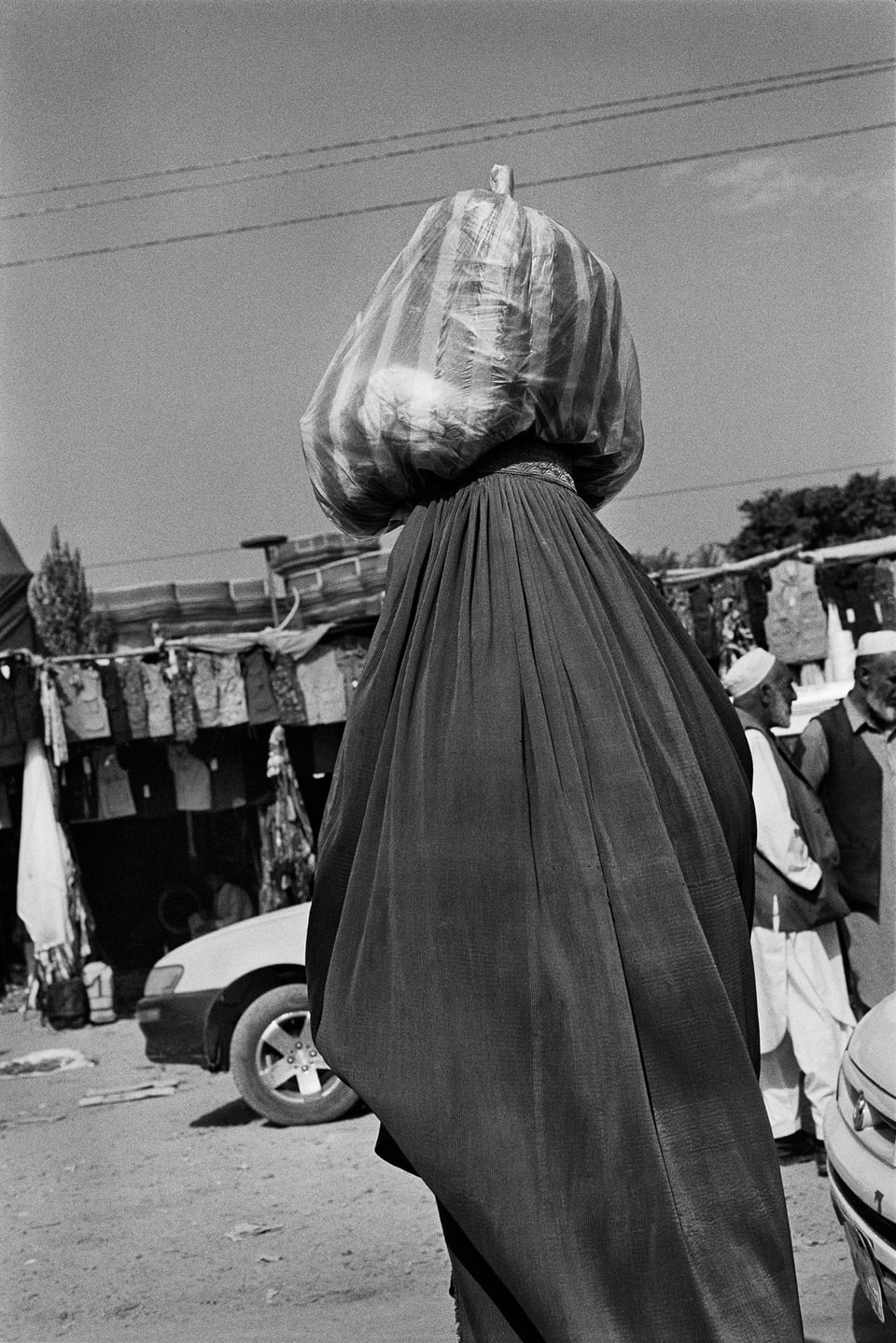 Black and white photo of a street scene. In the middle of the picture there is a person with a filled plastic bag on his head