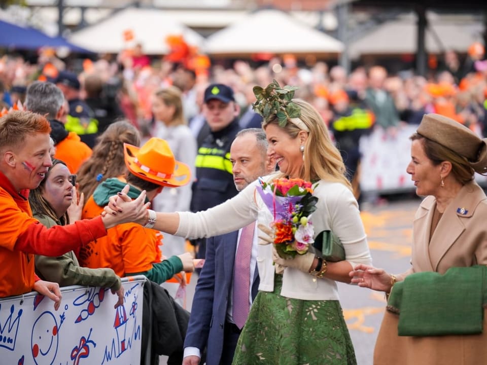 Frau in grünem Kleid nimmt Blumen von Menschenmenge entgegen während andere Person in braunem Mantel neben ihr steht.