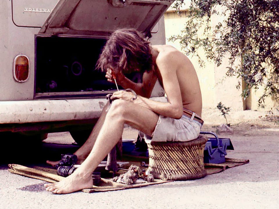 Ein junger Mann repariert vor seinem VW-Bus eine Zündkerze. Er hält einen Schraubenzieher in der Hand.