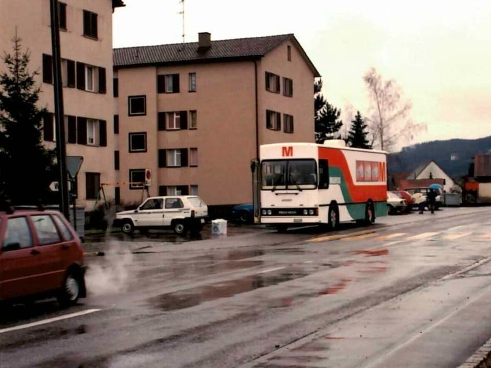Bus auf nasser Strasse in einer städtischen Umgebung mit Gebäuden und parkenden Autos.