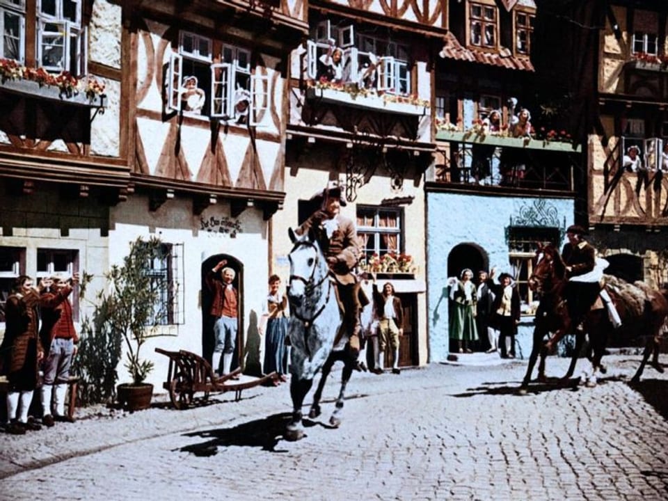 Filmszene mit Hans Albers als Münchhausen und Hermann Speelmans, die beide auf Pferden durch eine mittelalterliche Stadt reiten.