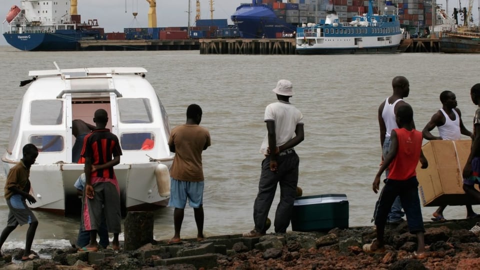 Männer aus Guinea-Bissau stehen am Ufer des Meeres und laden Kisten aus einem Motorboot.