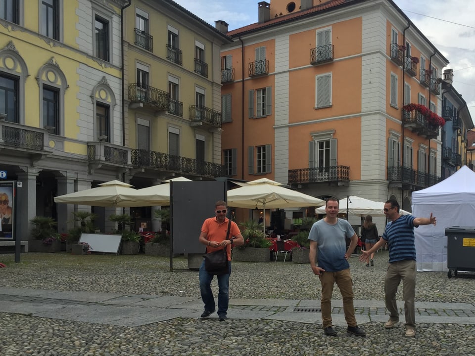 Platz in Locarno mit farbitgen, stattlichen Häusern. Im Vordergrund nähern sich schwungvoll drei Männer.