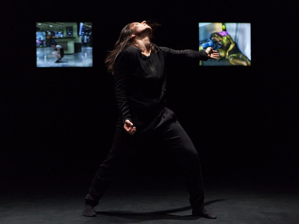 Eine Person in dunkler Kleidung tanzt vor dunklem Hintergrund, der mit drei Bildschirmen erleuchtet wird.