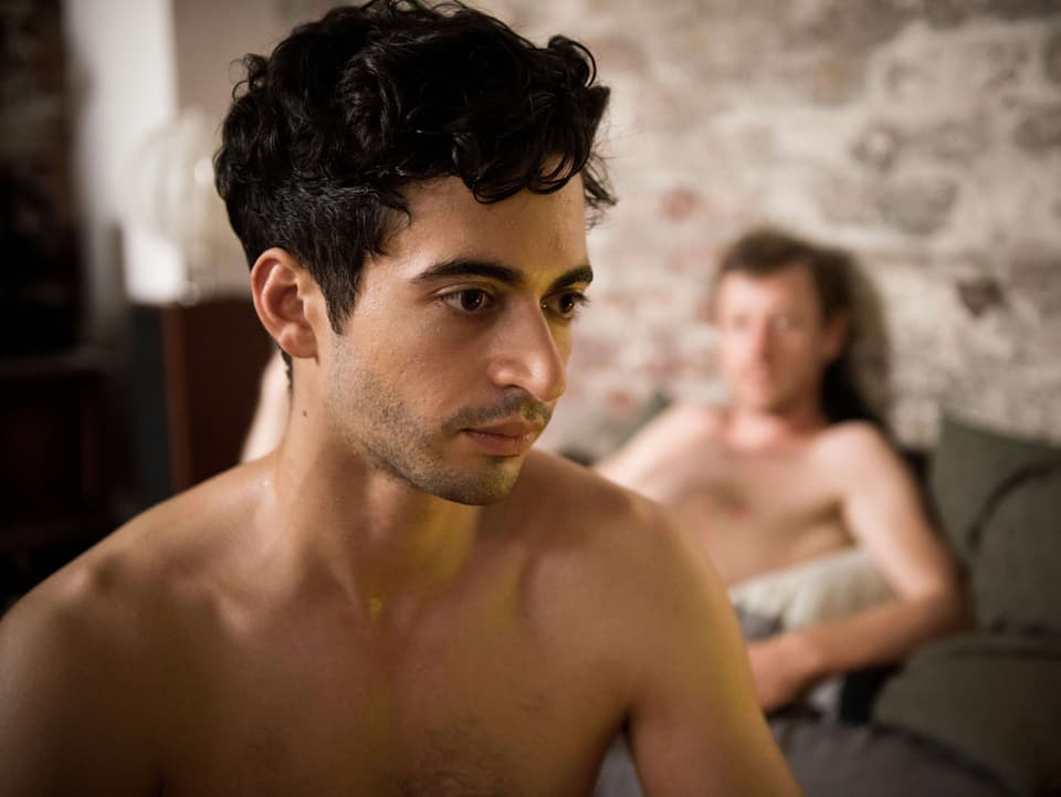 Filmszene ein junger Mann mit nacktem Oberkörper. Im Hintergrund liegt auf einem Bett ein nackter Mann.