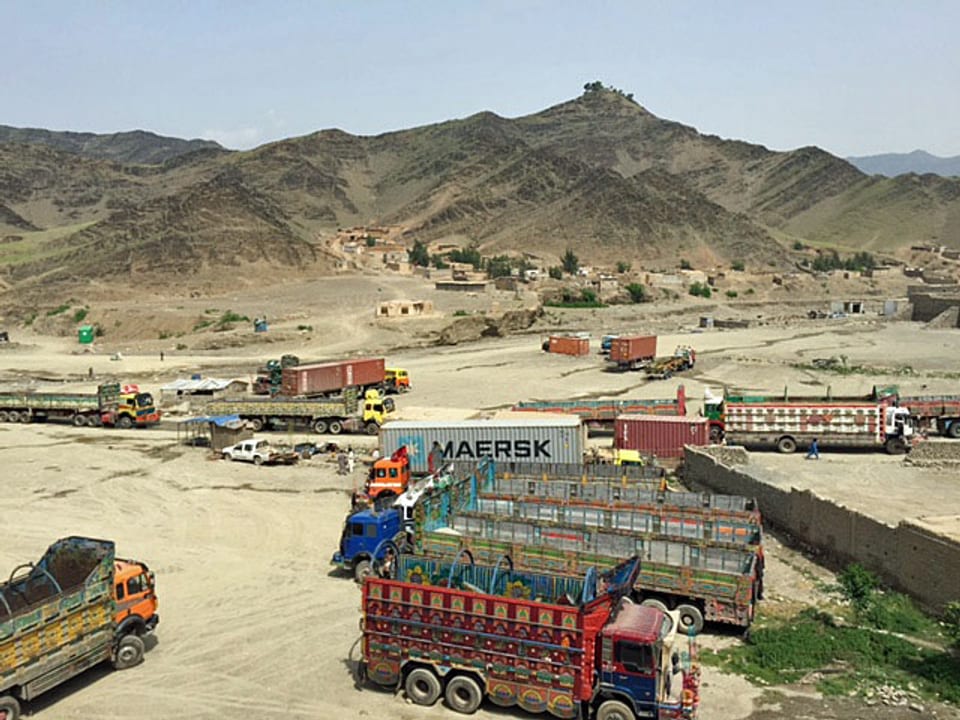 Viele Lastwagen stehen auf Wüstensand, hinten sind trockene Hügel zu sehen. Die Grenze ist fast nicht erkennbar, neu eine kleine Mauer ist zu sehen.