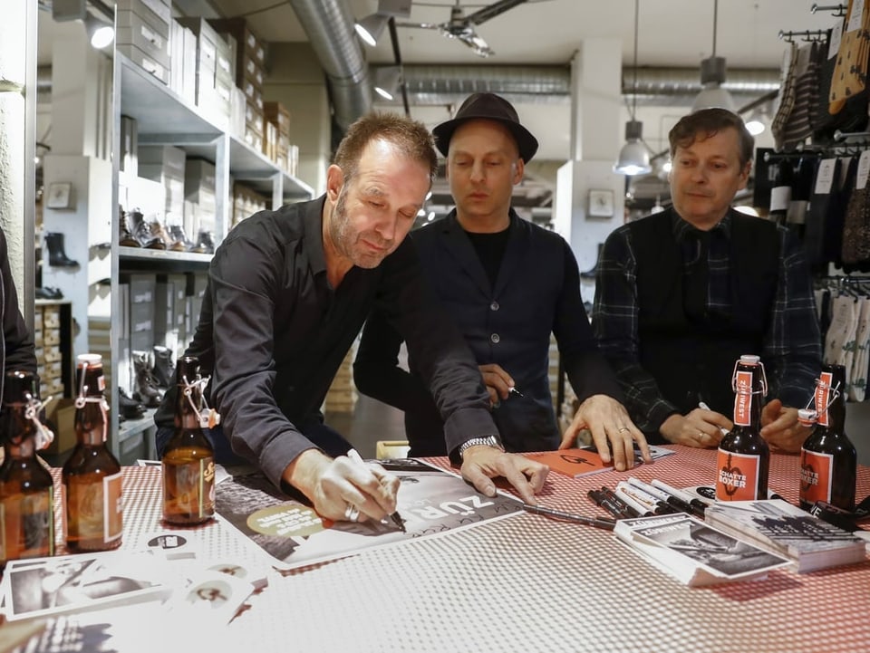Drei Männer stehen an einem Tisch, viele Biergläser stehen auch auf dem Tisch.