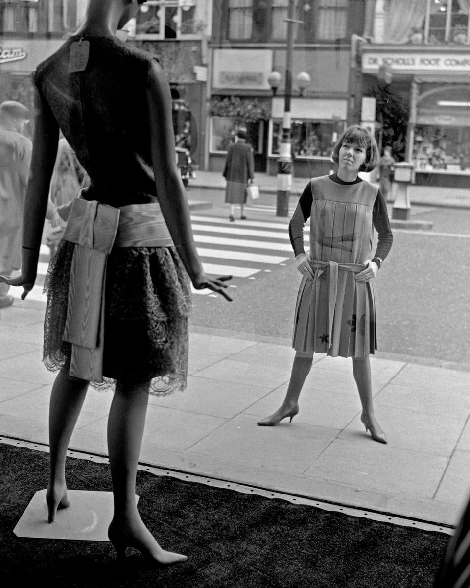 Schwarzweissfoto: Frau in Kleidung der 60er-Jahre schaut durch Schaufenster auf Mannequin