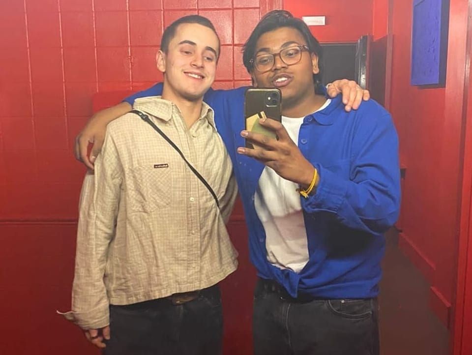 Justin steht mit einem Freund vor einem Spiegel, beide lachen und machen ein Selfie. 