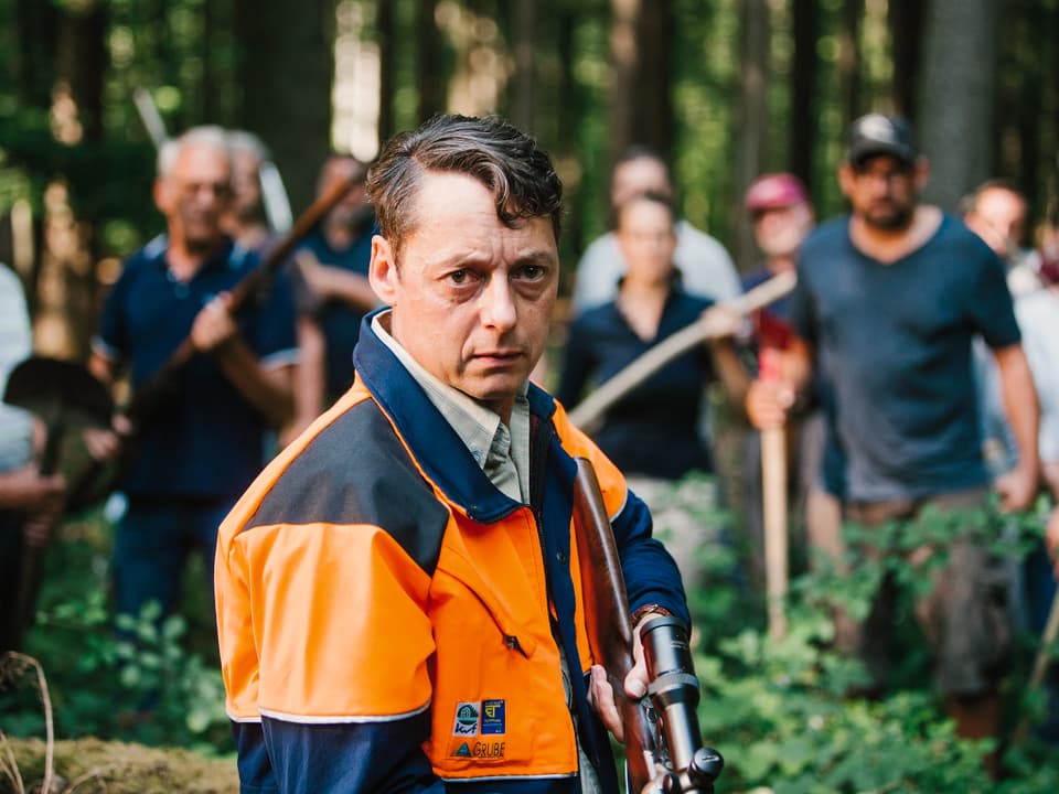 Ein Mann steht mit einem Gewehr im Wald. Hinter ihm sind bewaffenete Menschen