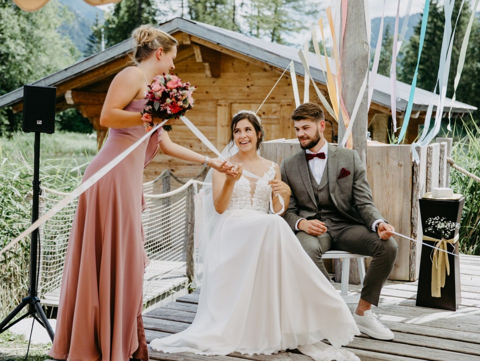 Ein Brautpaar sitzt vor einem kleinen Holzhaus, die Braut und eine Brautjunger halten sich bei der Hand.