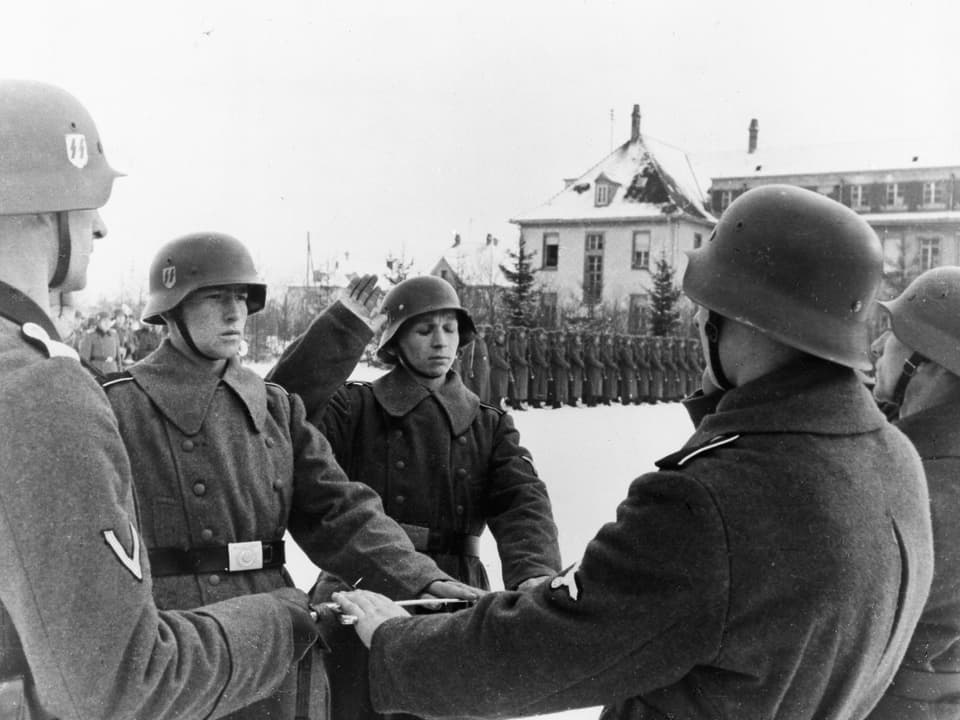 Schwarzweissfoto: Ein Mann in Uniform hebt die Hand zum Schwur. Er ist umgeben von weiteren Soldaten, im Hintergrund steht eine ganze Einheit Spalier.
