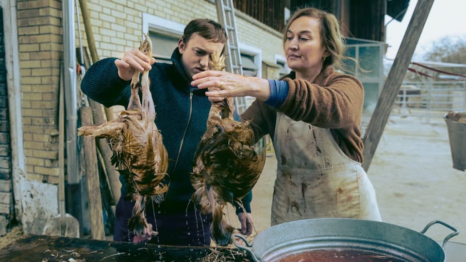 Frau und junger Mann halten je ein totes Huhn, vor ihnen ein Becken mit rotem Wasser.