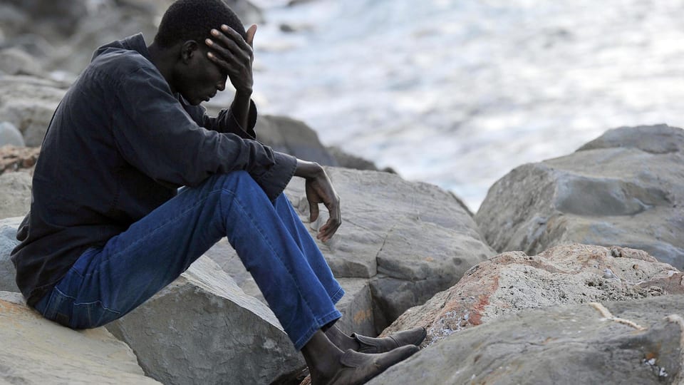 Ein Flüchtling sitzt auf einem Stein vor einem Meeres-Strand.