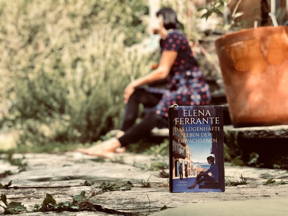 Der Roman «Das lügenhafte Leben der Erwachsenen» von Elena Ferrante steht im Vordergrund. Im Hintergrund sitzt Annette König auf einer Steintreppe.