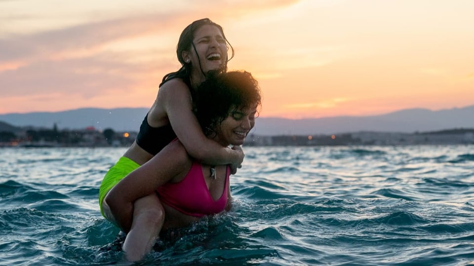 zwei Junge Frauen im Meer, eine Huckepack mit nassem Haar auf dem Rücken der anderen.