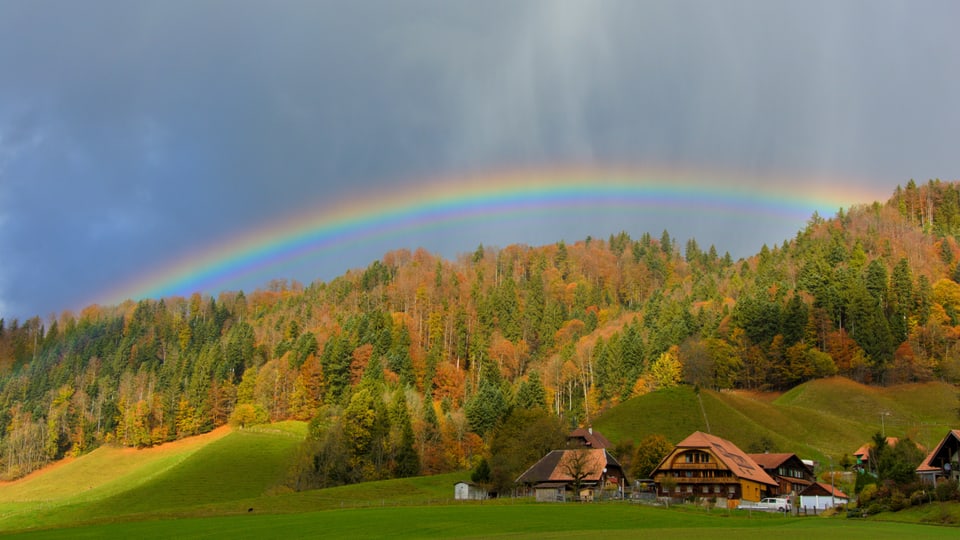 Regenbogen über herbstlich verfärbter Wald und Bauernhaus im Vordergrund.