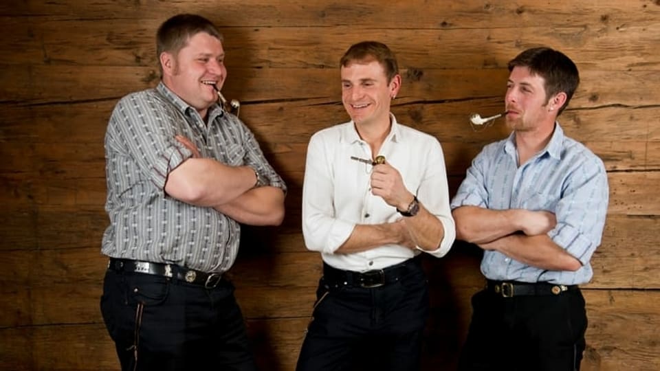 3 Männer lehnen lachend an eine Holzwand, jeder eine Tabakpfeife in der Hand oder im Mund. 