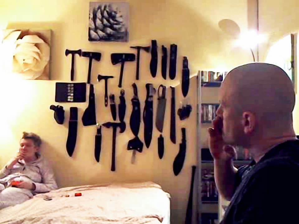 Blick durch eine Webcam: Ein Mann raucht im Vordergrund, im Hintergrund hängen Messer an der Wand.