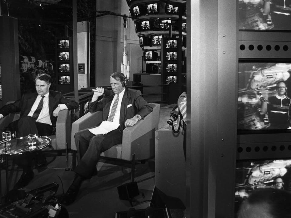 Schwarz-weiss Foto, Adolf Ogi und Mann sitzen in Raum umgeben von Bildschirmen, worauf Claude Nicollier ist.