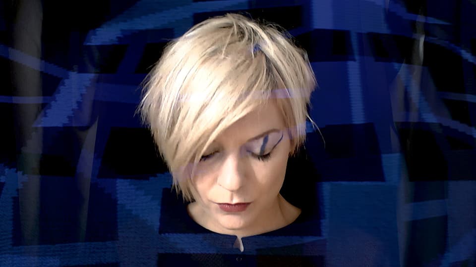 Eine blonde Frau mit kurzem Haar, deren blauschwarzes Kleid ihr über den Kopf zu wachsen scheint.