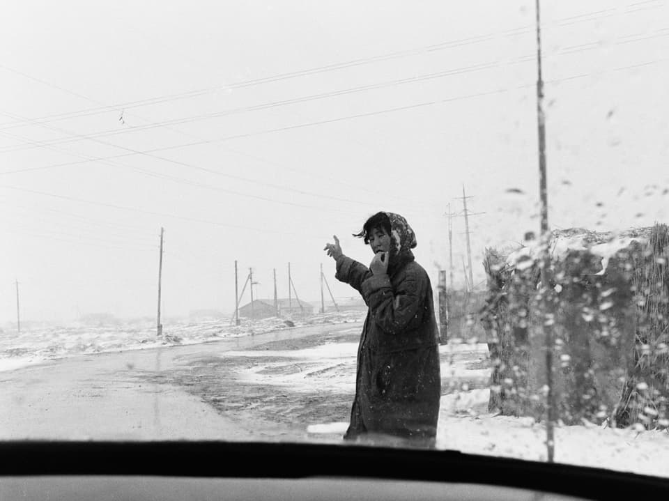 Eine Frau am Rand einer Landstrasse, die leicht mit Schnee bedeckt ist, aufgenommen durch eine Auto-Windschutzscheibe.