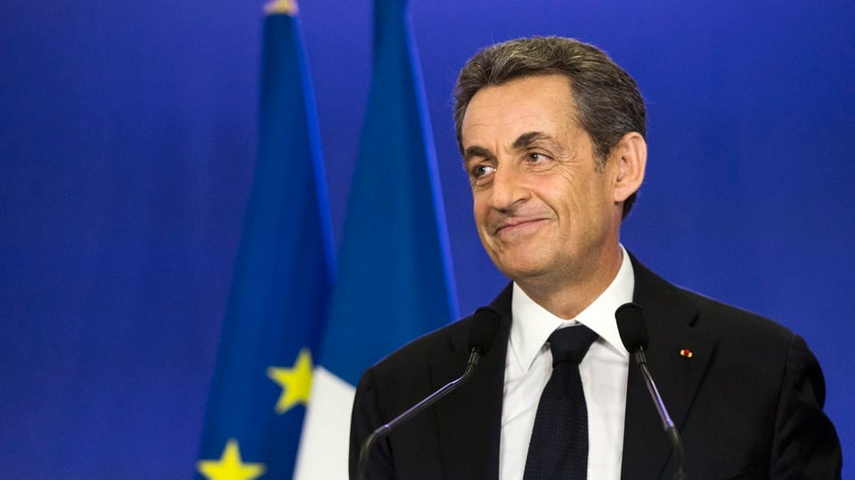Sarkozy lächelt verschmitzt. Im Hintergrund die französische und europäische Flaggen.