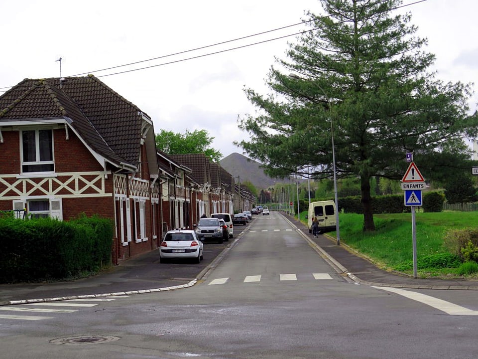 Aufnahme einer Quartierstrasse, auf der linken Strassenseite stehen Häuser mit einer dunkelroten Fassade