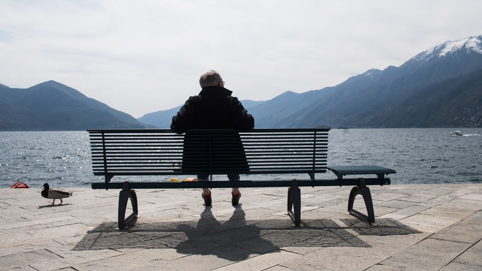 älterer Mann mit weissem Haar sitzt auf einer Bank, Rücken zur Kamera, Alpenpanorama im Blick.