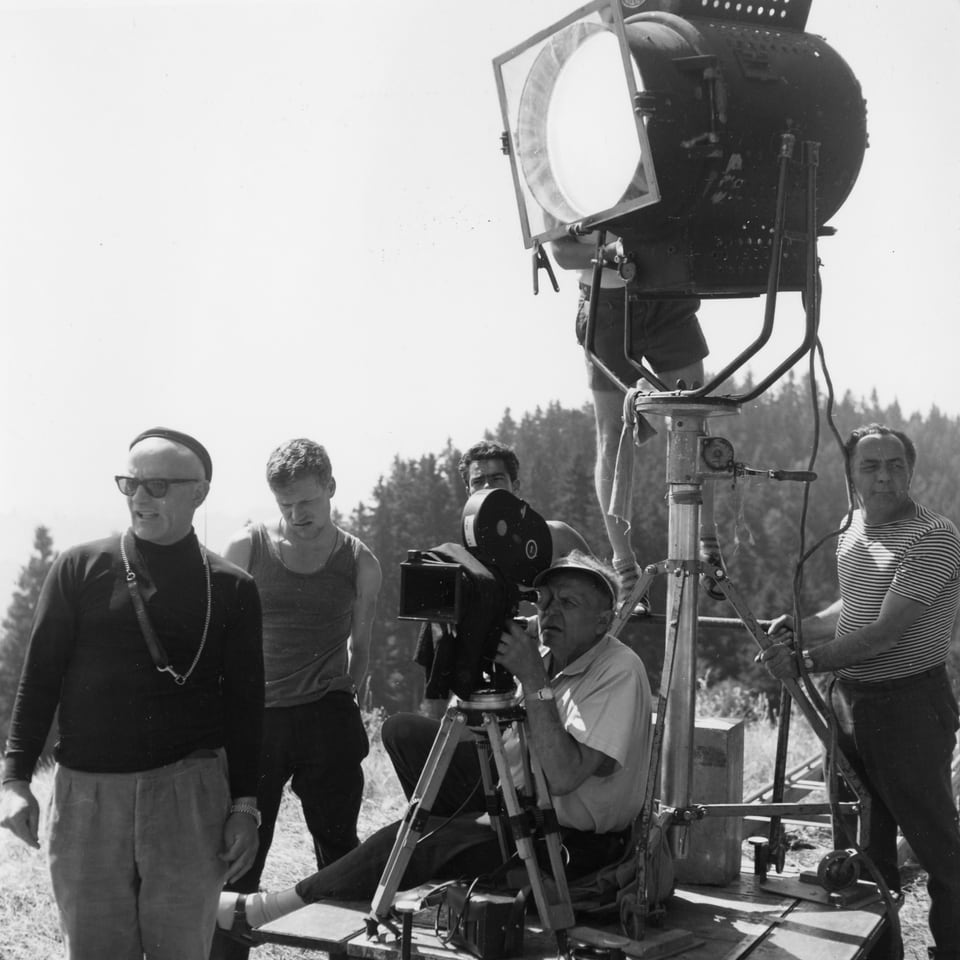 Ein grosser Filmscheinwerfer steht auf dem Kamerawagen auf einem Feld. Darunter die Kamera mit dem Kameramann. Franz Schnyder steht im Vordergrund.