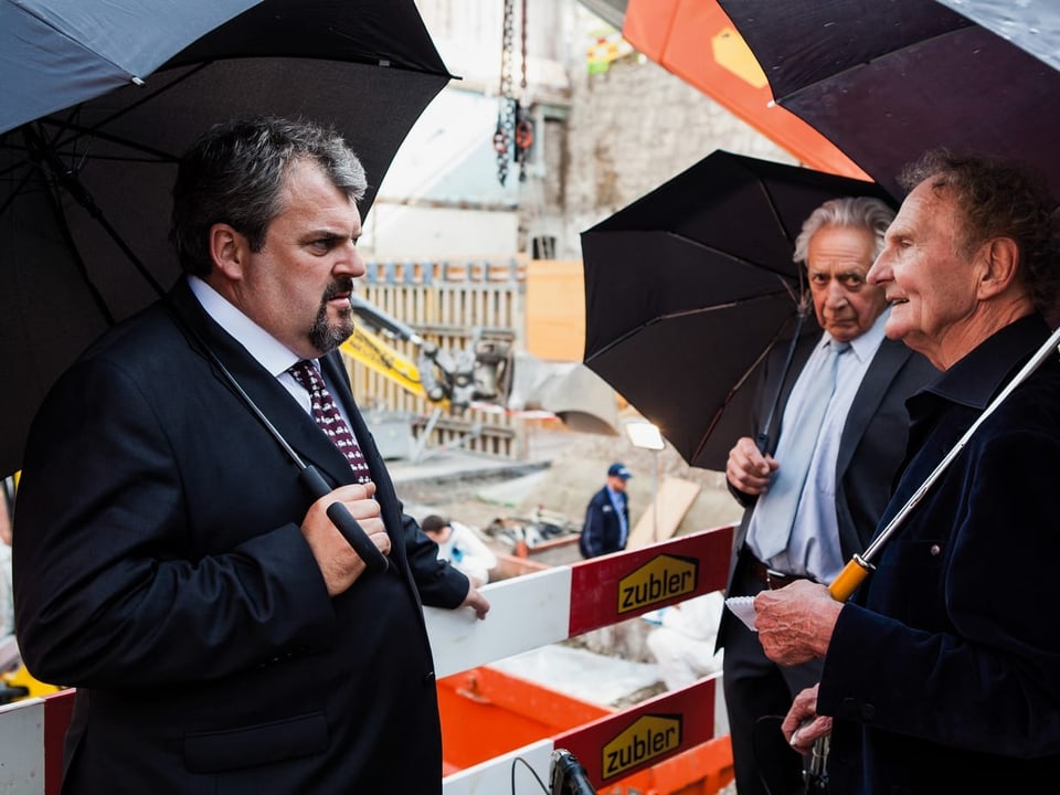 Drei Männer in Anzügen stehen unter Regenschirmen bei einer Baustelle