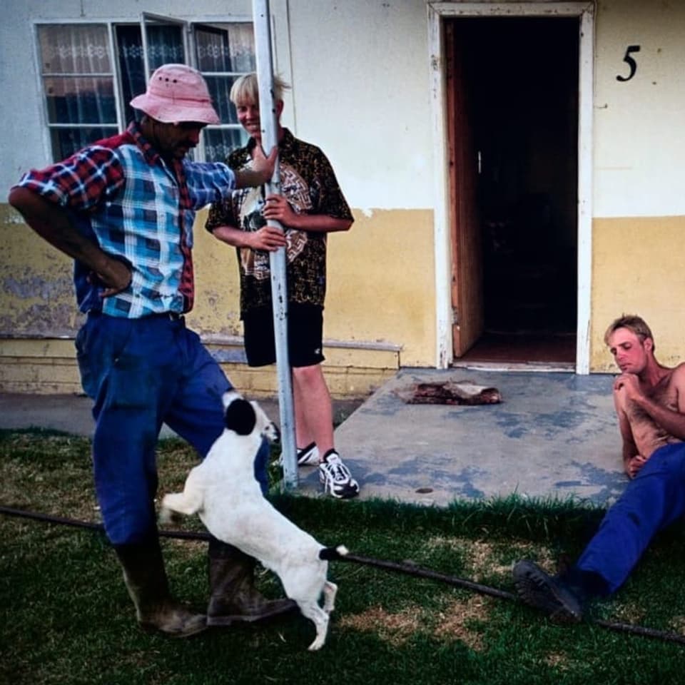 Ein schwarzer Mann im rosaroten Hut steht vor einem alten Haus und wird von vier weissen Männern angeschaut.