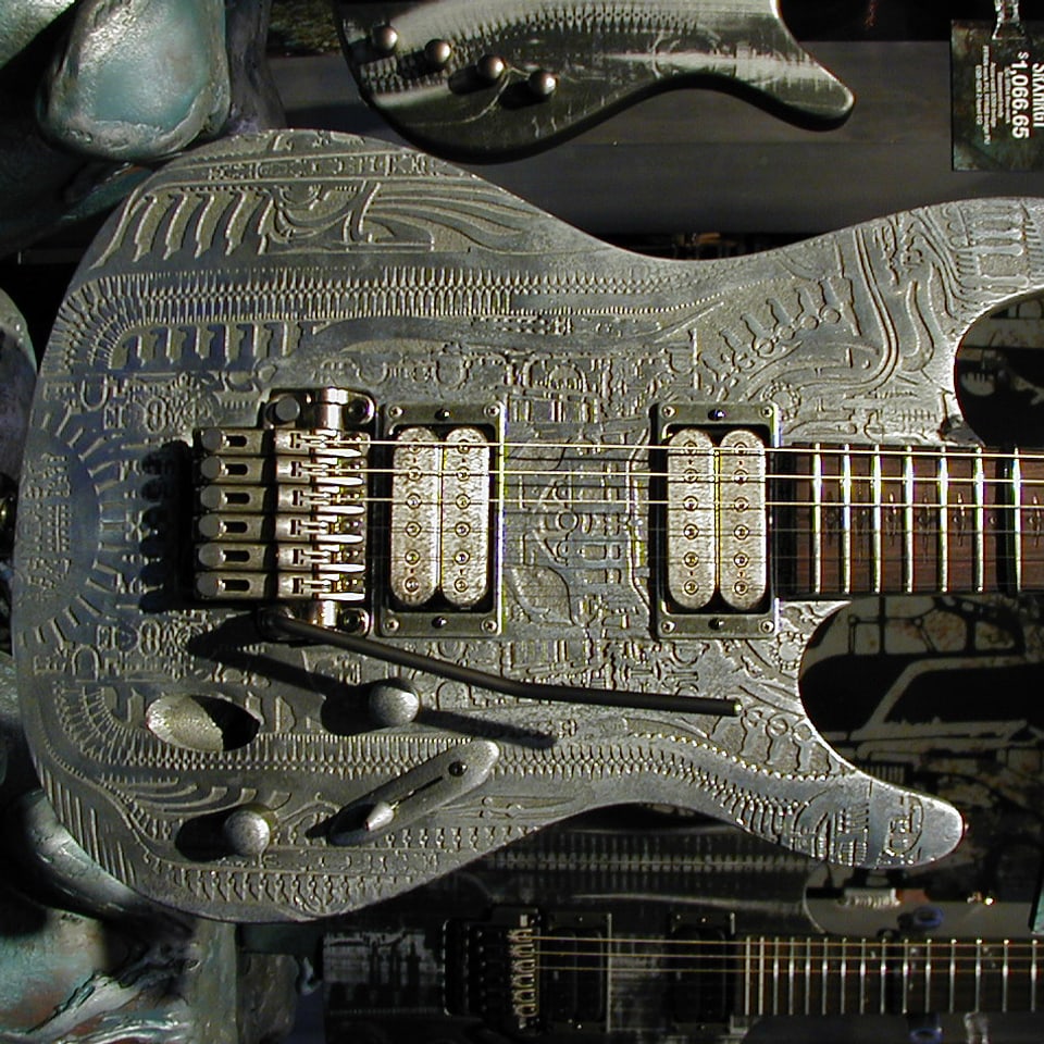 Zu sehen ist eine metallene Gitarre, die mit allerlei ornamentaler Kunst im Stile HR Gigers verziert ist.