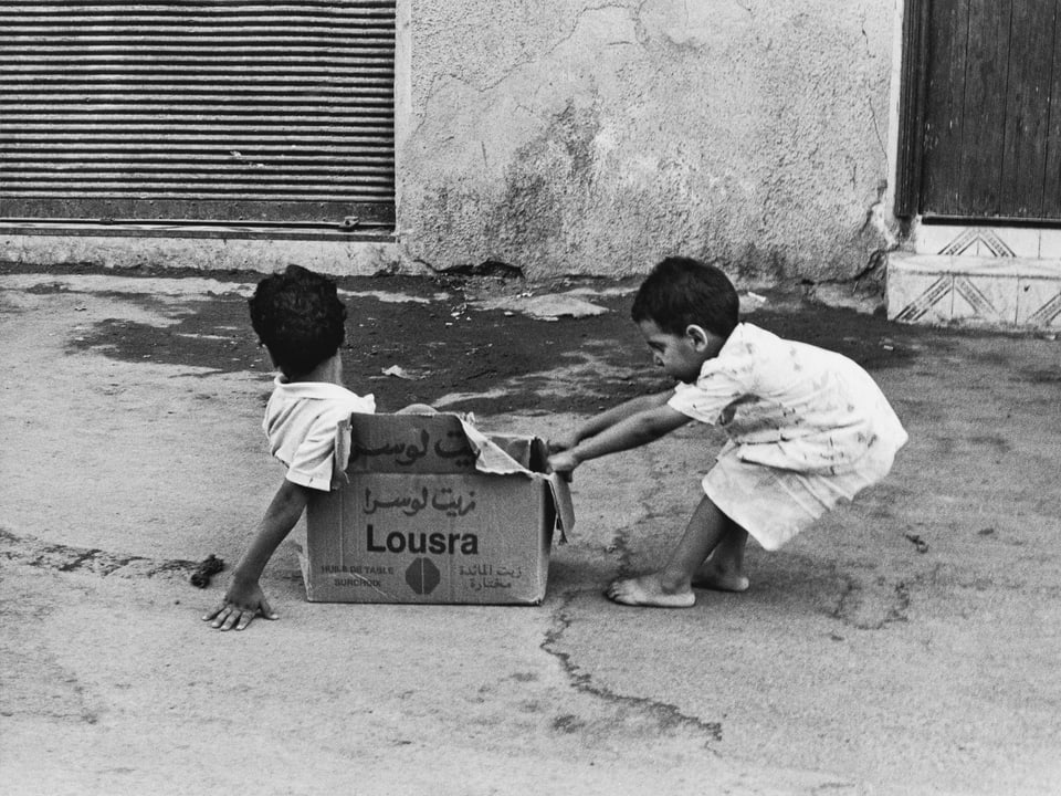 Schwarz-Weiss-Foto: Ein kleiner Junge zieht einen anderen Jungen in einer Kartonschachtel durch die Strasse. 