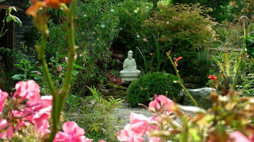 Ein Blumengarten mit Kiesweg und Buchsbäumen, in der Mitte des Bildes steht eine hüfthohe Buddha-Statue.