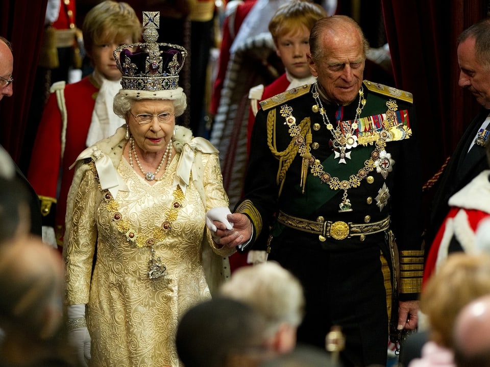 Alte Dame mit Krone hält die Hand eines älteren Herrn im Anzug.
