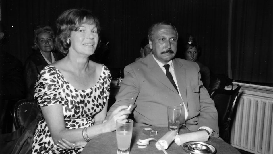 Ein Mann mit Schnauz neben einer rauchenden Frau.