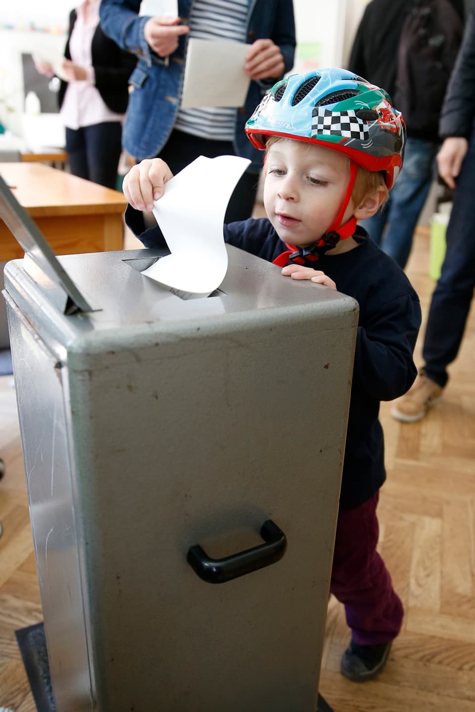Kind mit Velohelm steht vor einer Wahlurne und lässt einen Wahlzettel in den Schlitz fallen