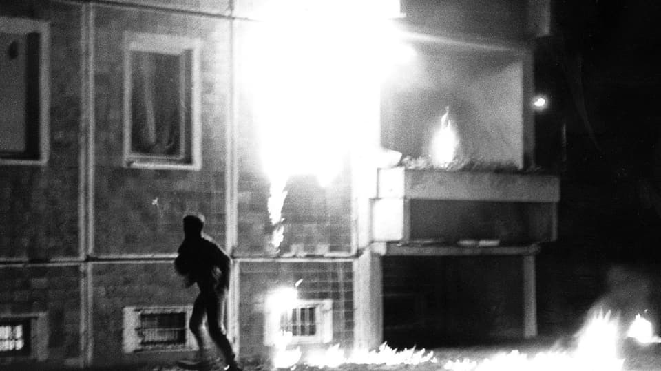 schwarzweiss Foto eines brennenden Gebäudes, davor rennt ein dunkler Umriss von rechts nach links aus dem Bild.