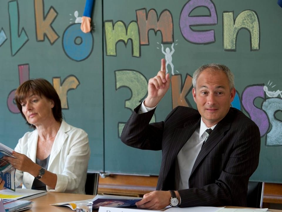 Bernhard Pulver hebt die Hand, hinter ihm eine Wandtafel mit dem Schriftzug «Willkommen in der 3. Klasse».
