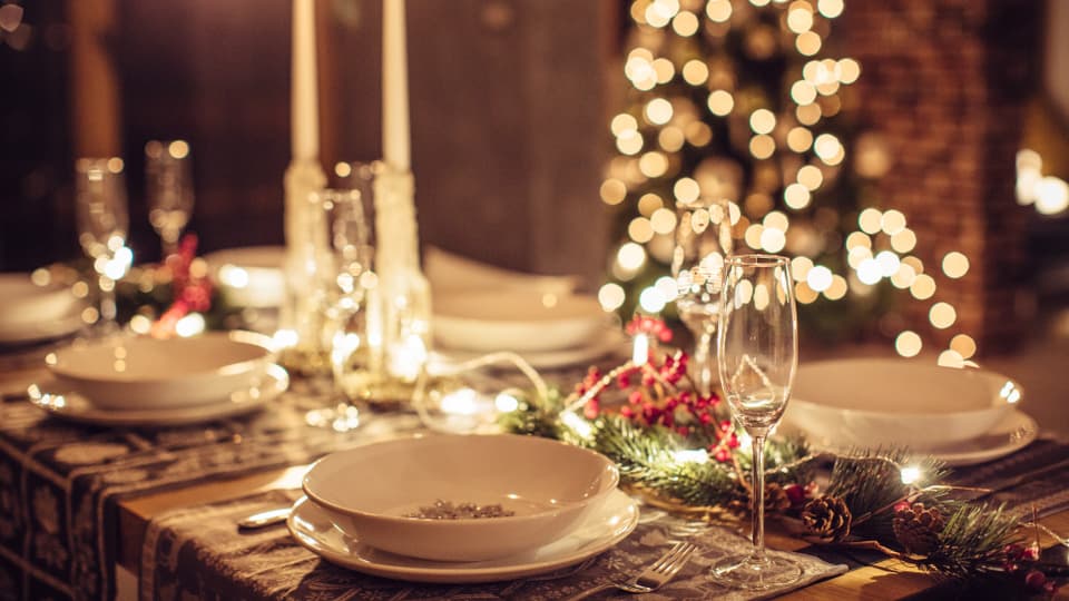 Weihnachtlich geschmückter und gedeckter Essenstisch mit mit einem beleuchteten Weihnachtsbaum im Hintergrund