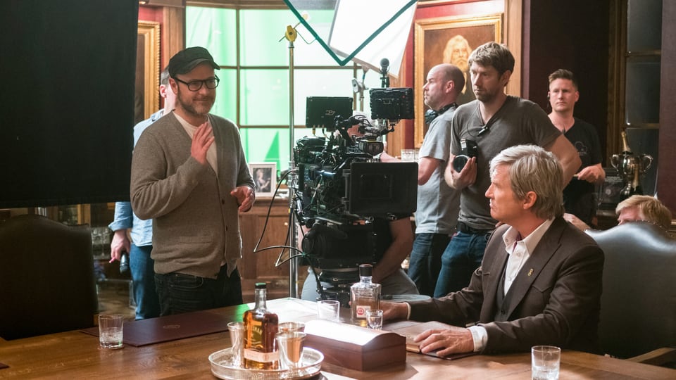 Filmset: Ein Mann sitzt am Tisch, ein Mann steht links von ihm, hat die Hand erhoben und scheint etwas zu erklären. Um die beiden herum stehen die Kameras und ein Film-Team.