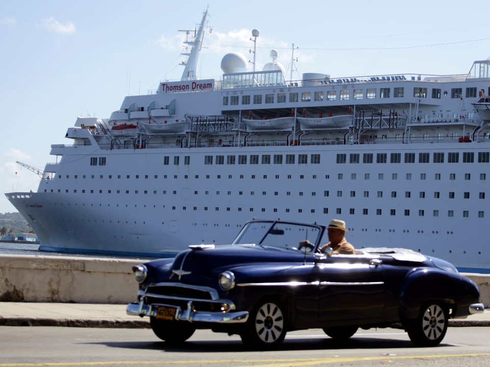 Ein Mann in seinem alten Wagen auf Kuba, dahinter liegt ein Kreuzfahrtschiff