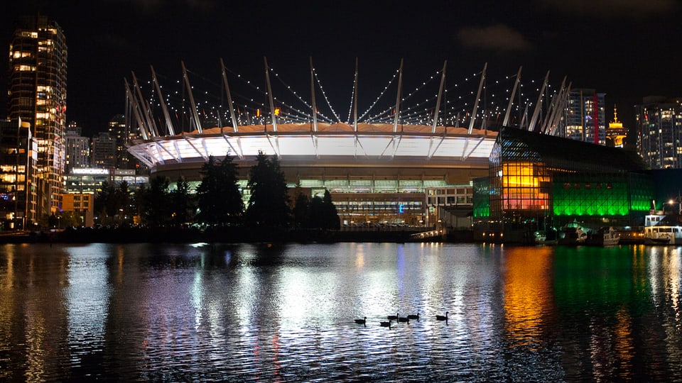 Blick auf das beleuchtete Stadion vom Wasser aus, eine Entenfamilie schwimmt vorbei.